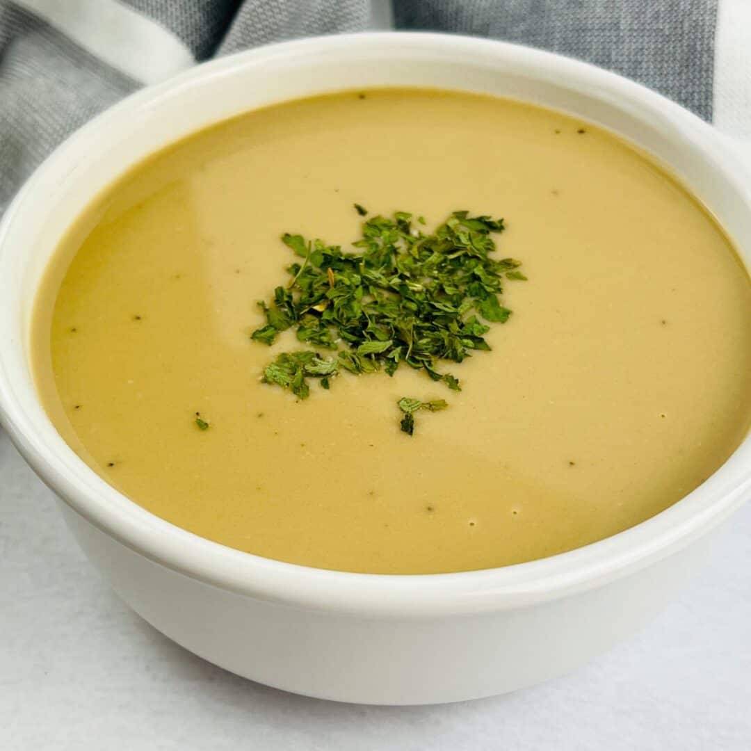 A white bowl filled with creamy brown ham gravy soup, garnished with chopped herbs, sits on a white surface with a gray and white cloth in the background.