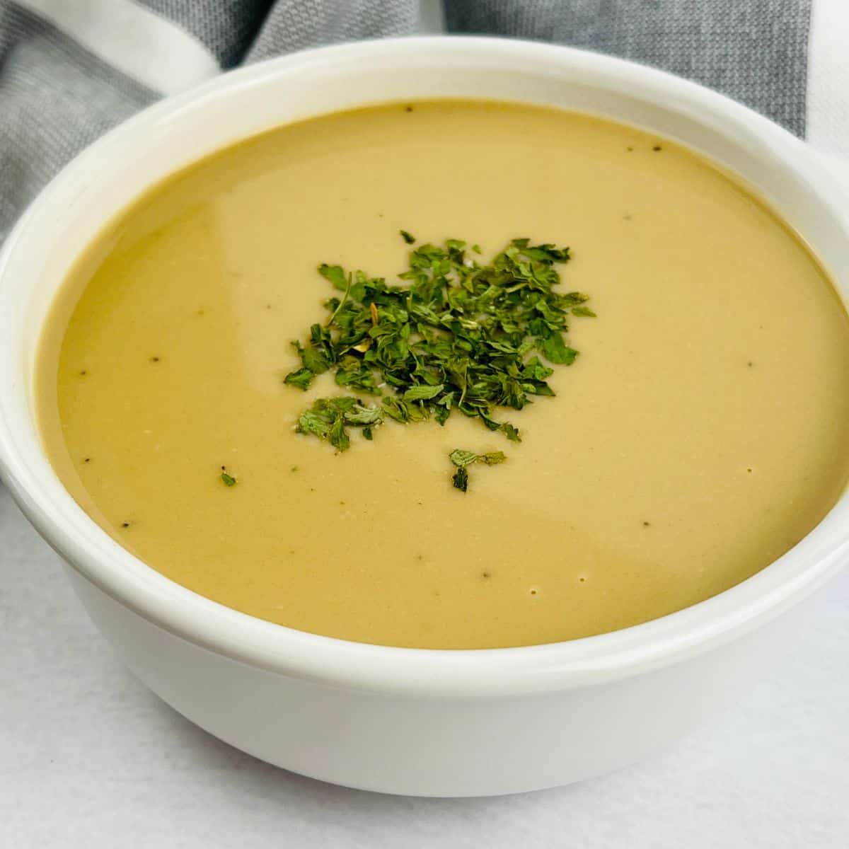A white bowl filled with creamy brown ham gravy soup, garnished with chopped herbs, sits on a white surface with a gray and white cloth in the background.
