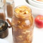 A glass jar of Homemade Apple Pie Filling sits on a white surface, surrounded by cinnamon sticks, apples, measuring cups, spice jars, and stacked white bowls.
