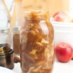 A glass jar of Homemade Apple Pie Filling sits on a white counter, surrounded by fresh apples, cinnamon sticks, measuring cups, and other kitchen items.