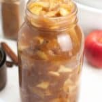 A glass jar filled with Homemade Apple Pie Filling, surrounded by cinnamon sticks and fresh apples in the background.