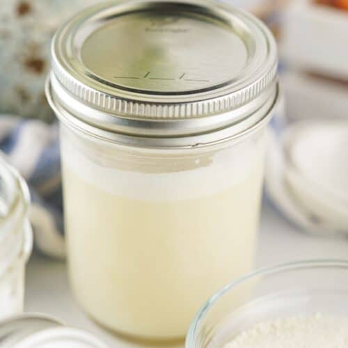 A glass jar filled with a creamy liquid sits on a white surface next to a small bowl of powder and another jar, perfectly capturing the essence of a homemade Sweetened Condensed Milk Substitute.