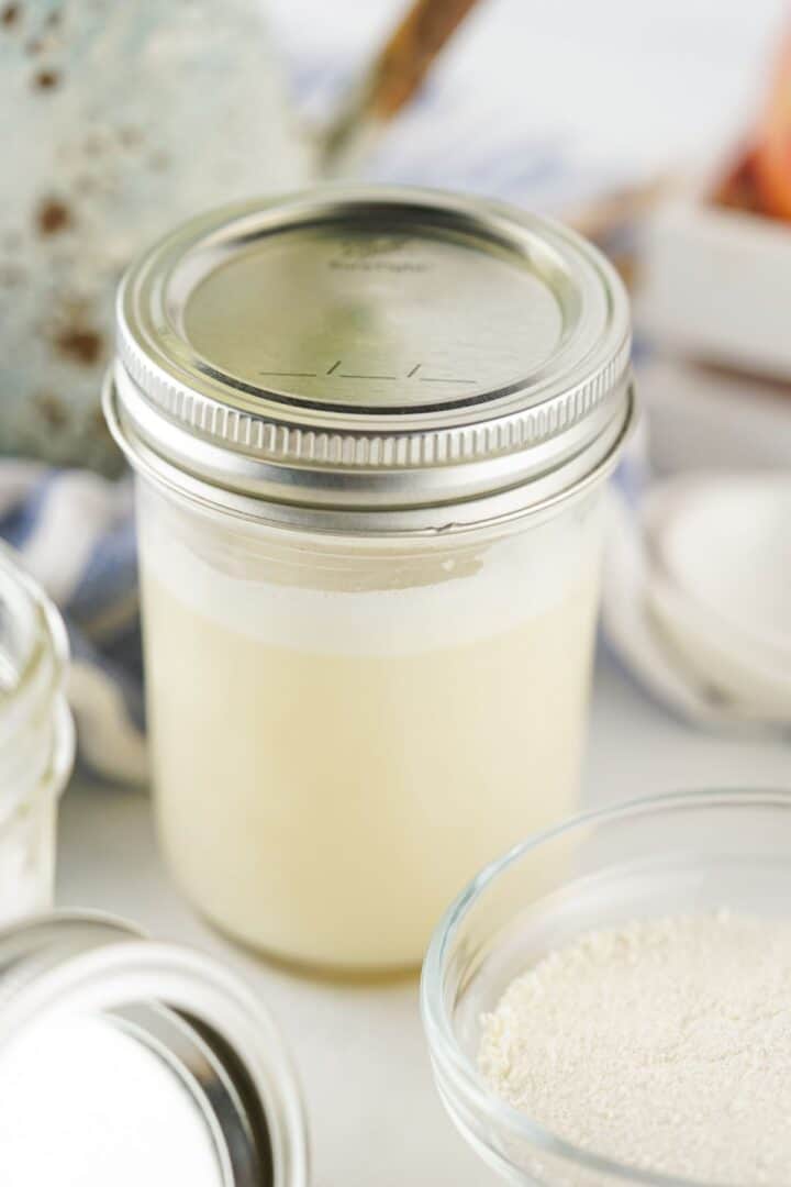 A glass jar filled with a creamy liquid sits on a white surface next to a small bowl of powder and another jar, perfectly capturing the essence of a homemade Sweetened Condensed Milk Substitute.