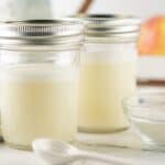Two glass jars filled with a white liquid, likely a sweetened condensed milk substitute or yogurt, sit on a counter next to bowls of white powder and a striped cloth, with apples blurred in the background.