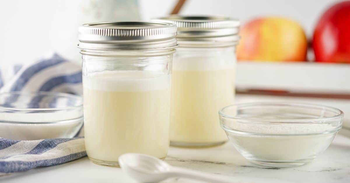 Two glass jars filled with a white liquid, likely a sweetened condensed milk substitute or yogurt, sit on a counter next to bowls of white powder and a striped cloth, with apples blurred in the background.