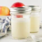 Two glass jars filled with pale milk substitute, topped with lids, set on a white surface with apples and a striped cloth in the background.