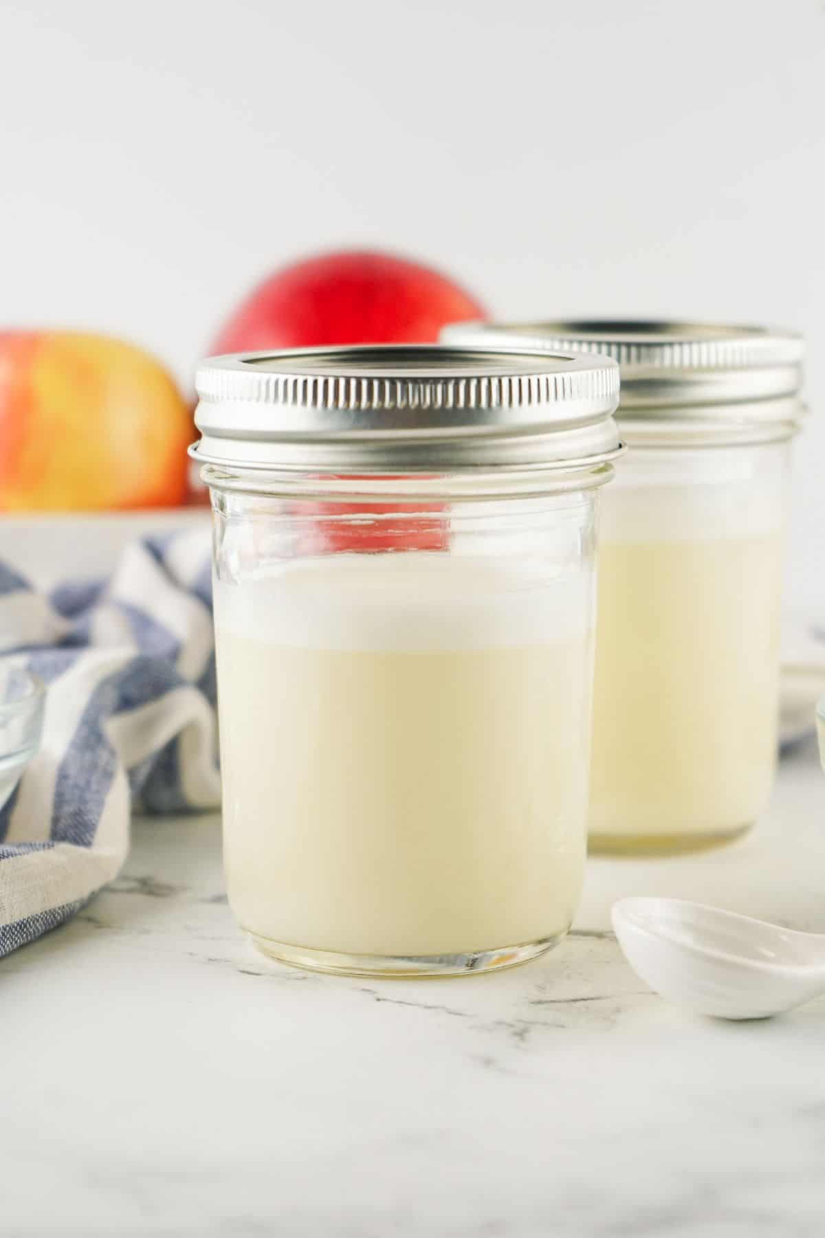 Two glass jars filled with pale  milk substitute, topped with lids, set on a white surface with apples and a striped cloth in the background.