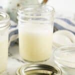 A glass jar filled with a creamy Sweetened Condensed Milk Substitute sits on a marble surface beside a metal lid, a bowl of powder, and a striped cloth in the background.