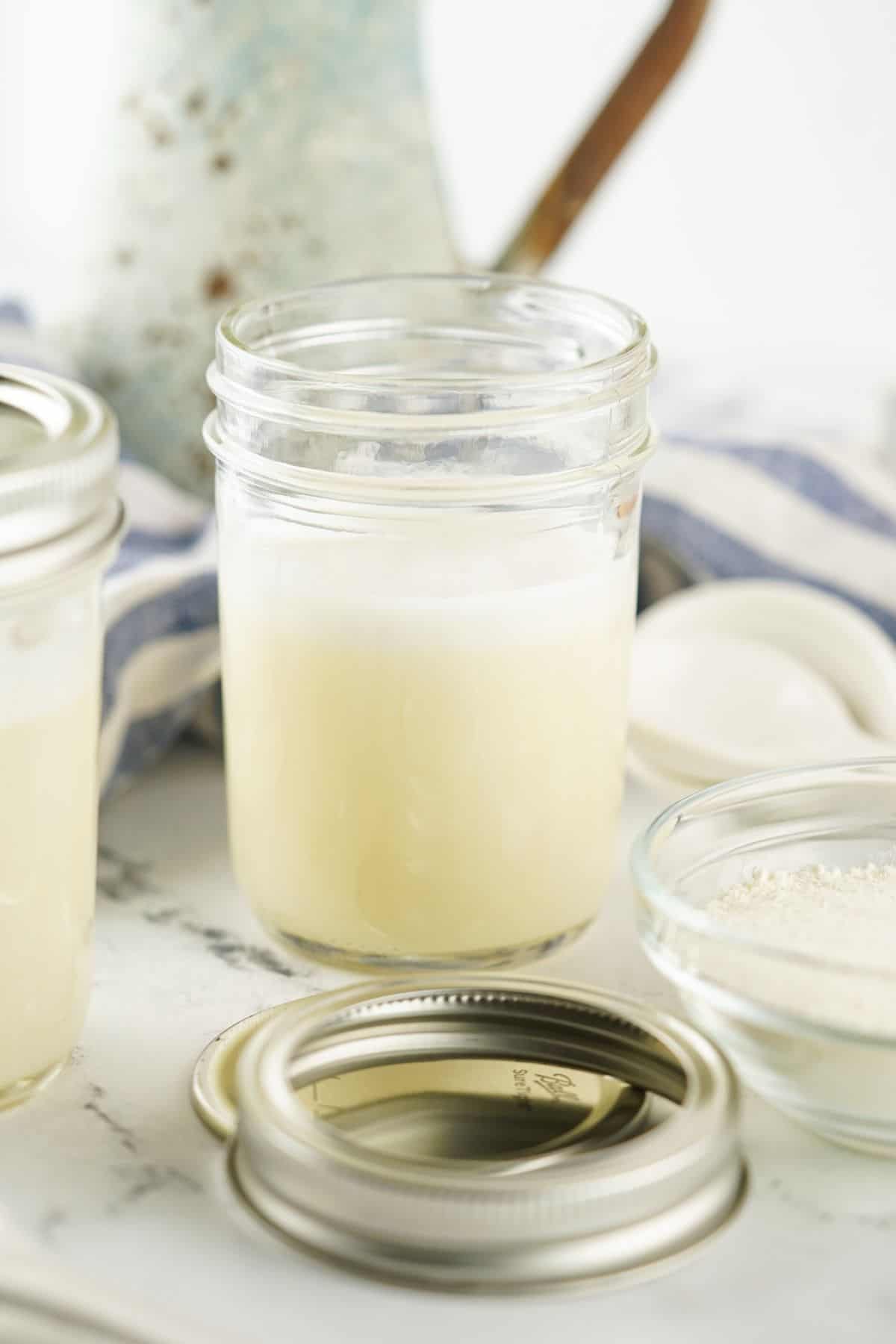 A glass jar filled with a creamy Sweetened Condensed Milk Substitute sits on a marble surface beside a metal lid, a bowl of powder, and a striped cloth in the background.