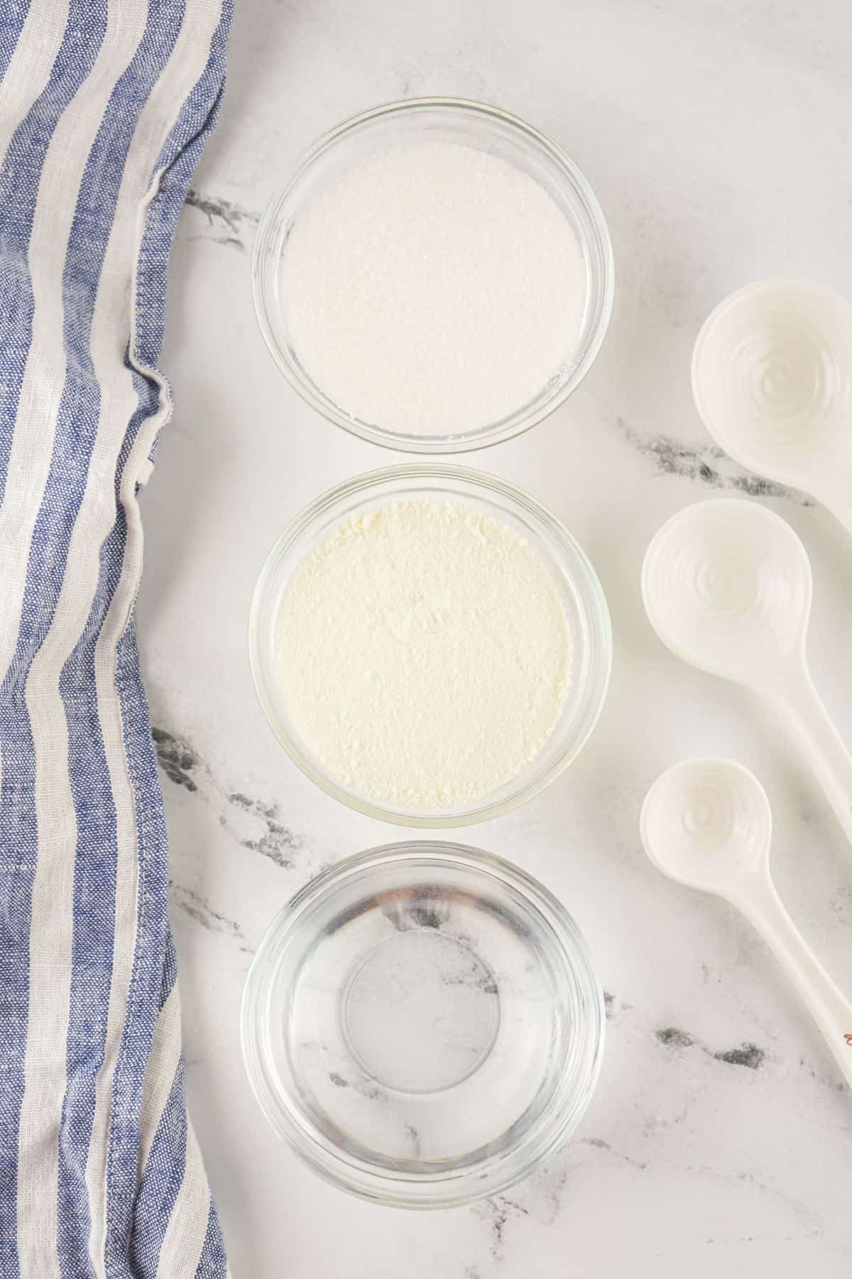 Three glass bowls filled with sugar, powdered milk, and water—perfect for making a Sweetened Condensed Milk Substitute—are arranged vertically next to white measuring spoons and a blue striped cloth on a marble surface.