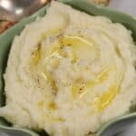 A bowl of Instant Pot Mashed Potatoes topped with melted butter and a sprinkle of black pepper, with a spoon and decorative napkin in the background.