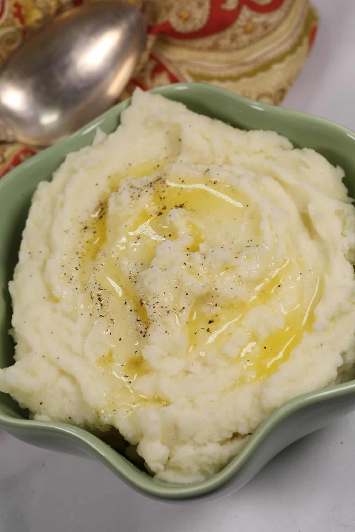 A bowl of Instant Pot Mashed Potatoes topped with melted butter and a sprinkle of black pepper, with a spoon and decorative napkin in the background.