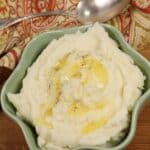 A bowl of creamy Instant Pot Mashed Potatoes topped with melted butter and black pepper, placed on a wooden surface beside a patterned cloth and a spoon.
