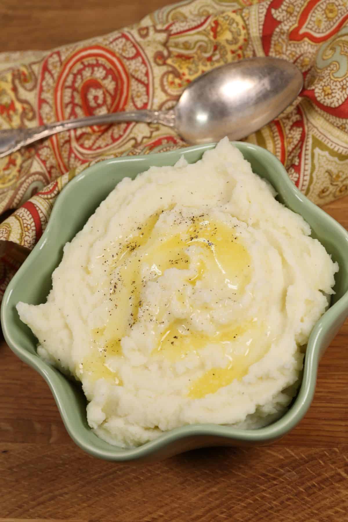A bowl of creamy Instant Pot Mashed Potatoes topped with melted butter and black pepper, placed on a wooden surface beside a patterned cloth and a spoon.