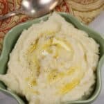 A bowl of Instant Pot Mashed Potatoes topped with melted butter and black pepper sits beside a vintage spoon and a patterned cloth napkin.