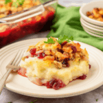 A serving of Thanksgiving Leftovers Casserole with mashed potatoes, stuffing, cranberry sauce, and herbs on a plate, a fork beside it, and more casserole in the background.