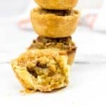 Four mini pecan pies, also known as pecan tarts, are stacked on a white surface, with the front pie cut open to reveal the rich, nutty filling inside.