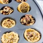 A muffin tin containing baked mini Pecan Tarts, some filled with chopped pecans and some partially empty.