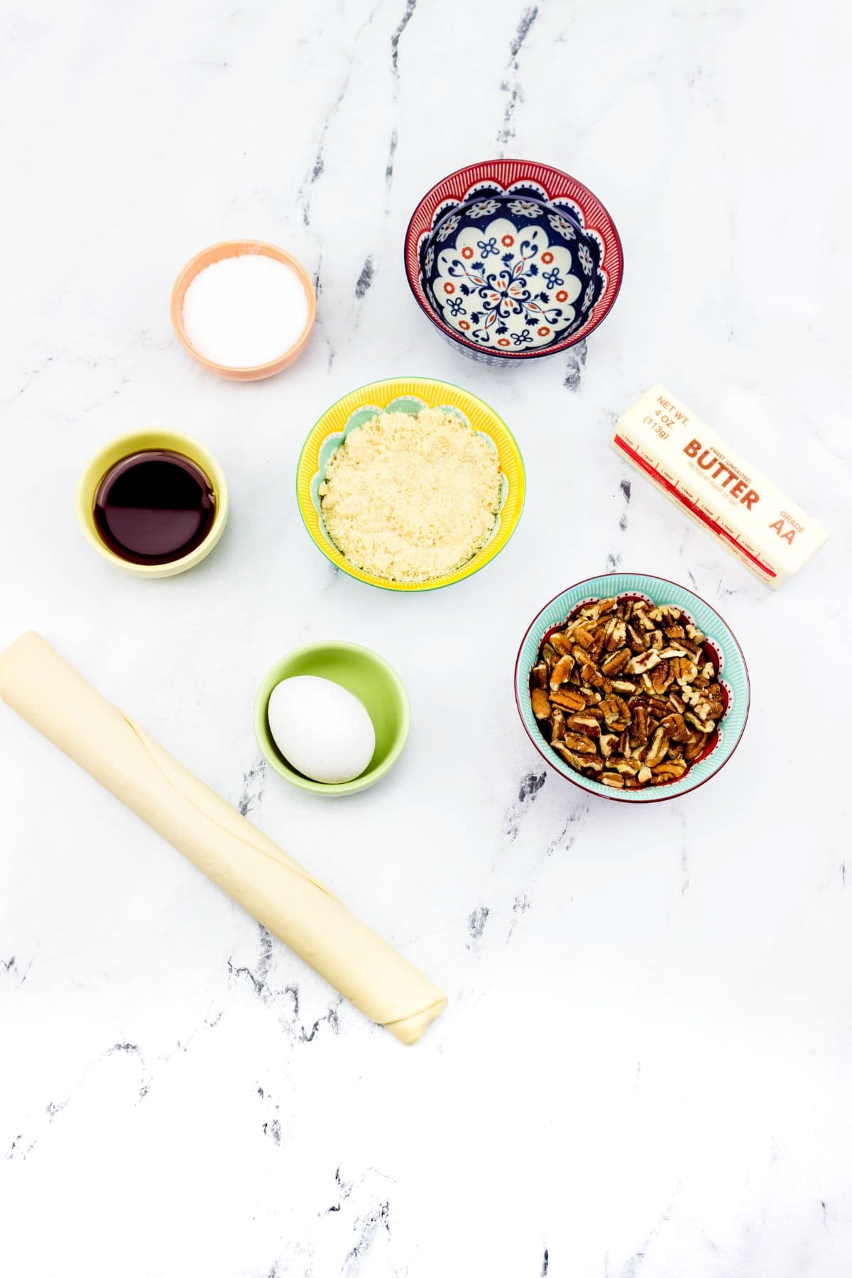 Ingredients  arranged on a white surface, including sugar, butter, chopped pecans, an egg, vanilla extract, a pastry sheet, and two other small bowls with dry ingredients.