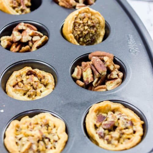 A muffin tin holds several mini pecan pies and pecan tarts, some baked and some filled with raw pecan pieces, on a marble countertop.