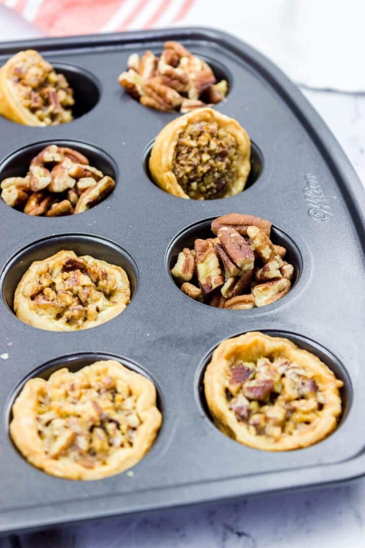 A muffin tin holds several mini pecan pies and pecan tarts, some baked and some filled with raw pecan pieces, on a marble countertop.