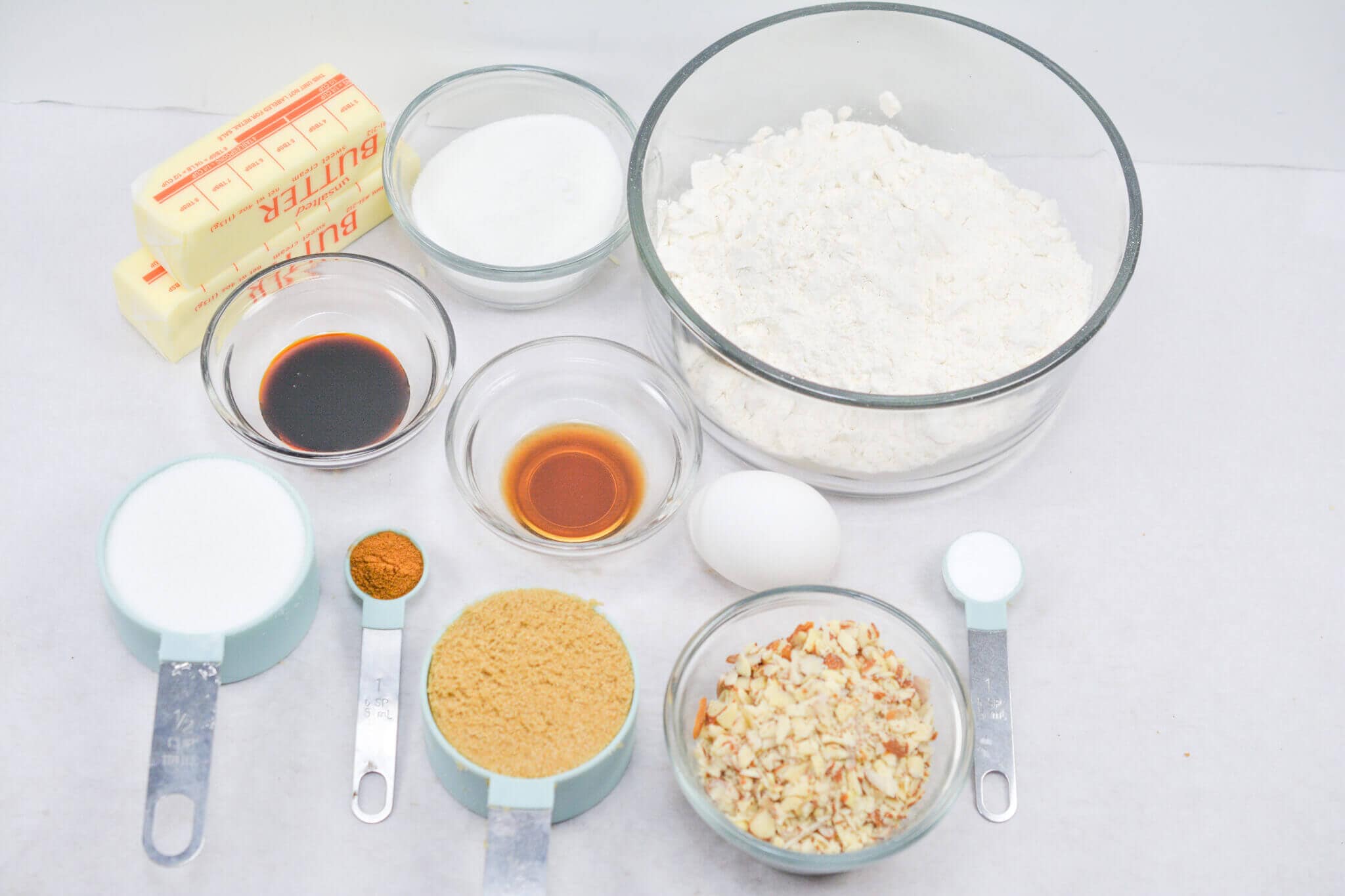 A delightful assortment for Norwegian cookies awaits: butter, flour, sugar, brown sugar, vanilla extract, almond extract, and an egg blend together alongside fragrant cinnamon and chopped almonds. Measuring spoons rest on the pristine white surface as you prepare this traditional treat.