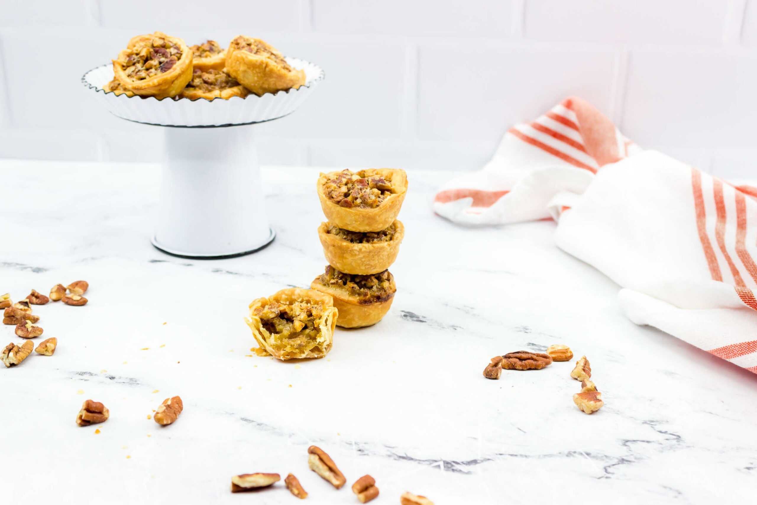 Mini pecan pies and Pecan Tarts are stacked on a marble surface, with more pies on a cake stand in the background. Pecans and a white and red-striped towel are scattered nearby, adding to the inviting display.