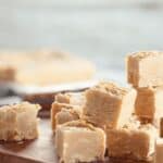 A stack of light brown Penuche Fudge squares is arranged on a wooden board, with more fudge pieces blurred in the background.