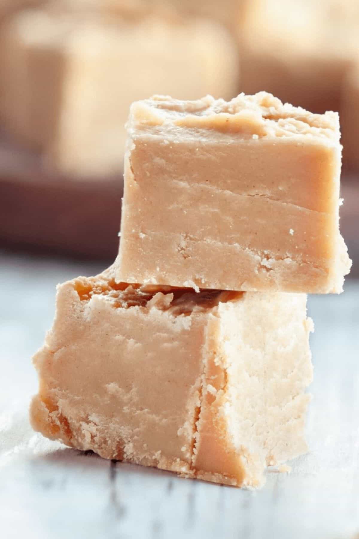 Two pieces of light brown, square-shaped penuche fudge are stacked on top of each other on a light surface, with more fudge pieces blurred in the background.
