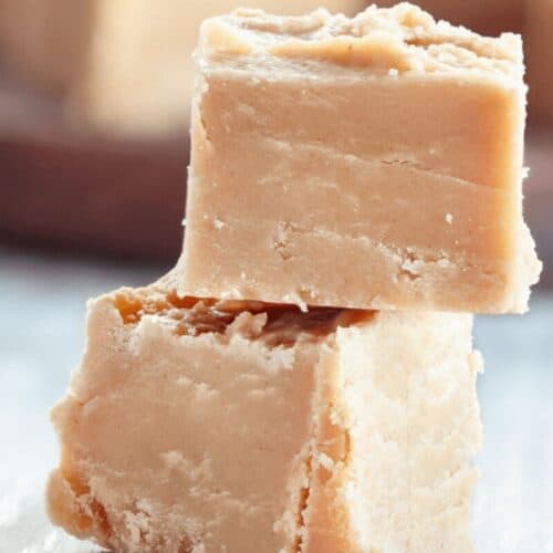 Two pieces of light brown penuche fudge are stacked on top of each other on a light surface, with more pieces blurred in the background.
