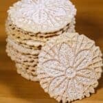 A stack of round, patterned Pizzelle Cookies dusted with powdered sugar sits on a wooden surface, with one cookie leaning against the stack.