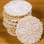 A stack of round, patterned Pizzelle Cookies dusted with powdered sugar sits on a wooden surface, with one cookie propped in front.