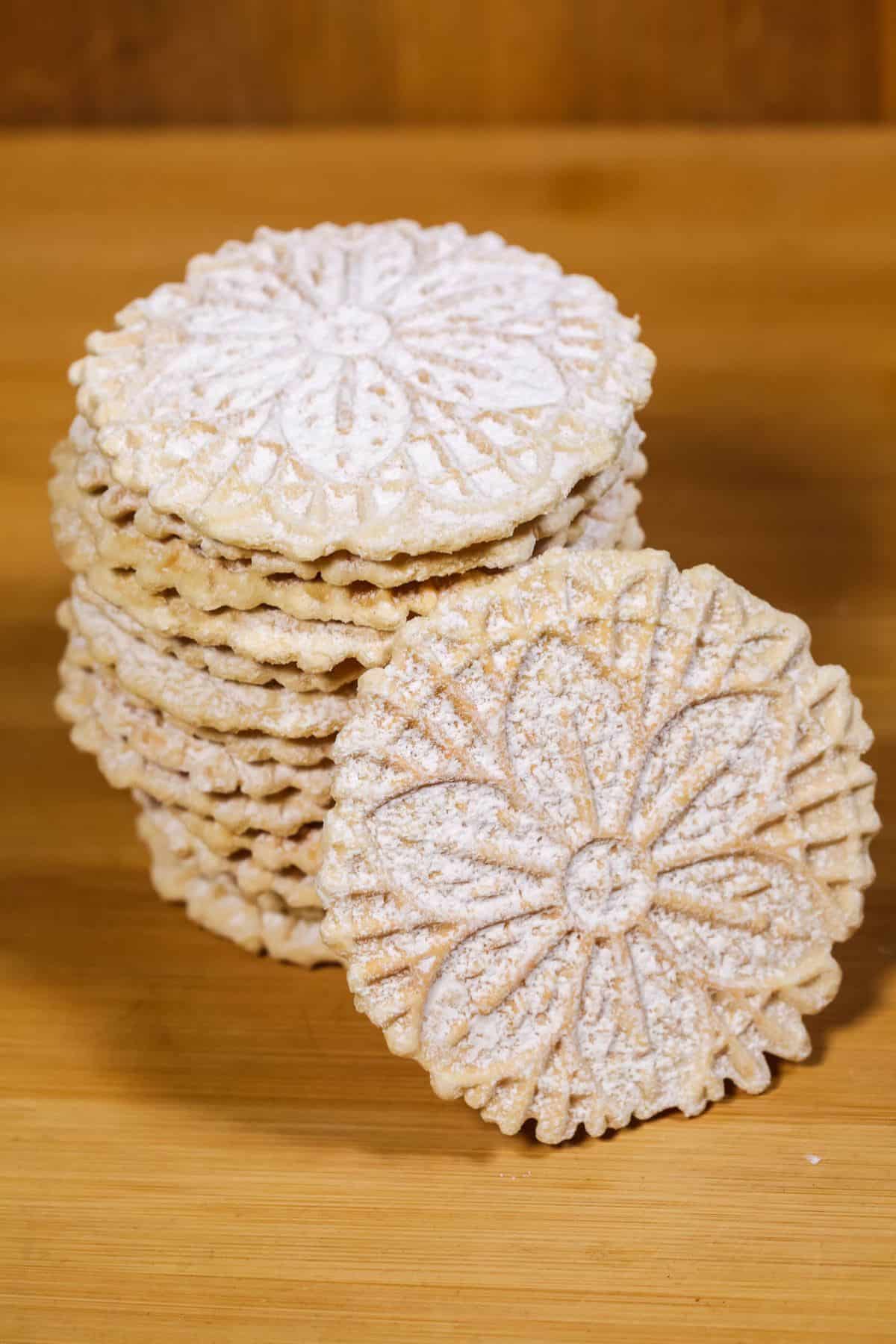A stack of round, patterned Pizzelle Cookies dusted with powdered sugar sits on a wooden surface, with one cookie propped in front.