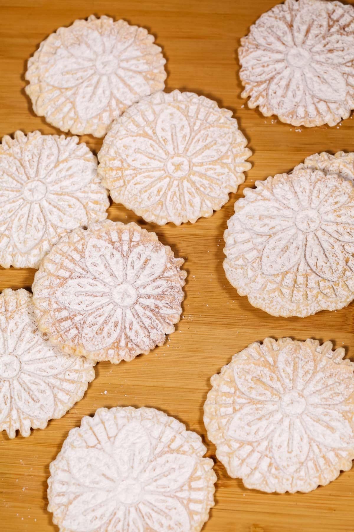 Pizzelle cookies with intricate floral patterns, dusted with powdered sugar, are beautifully arranged on a wooden surface.
