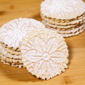 Several stacks of round, floral-patterned Pizzelle Cookies dusted with powdered sugar are arranged on a wooden surface.