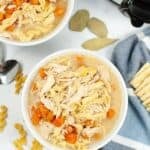 Two bowls of savory Slow Cooker Turkey Noodle Soup with carrots and noodles sit on a white surface near a slow cooker, bay leaves, a spoon, crackers, and uncooked pasta.