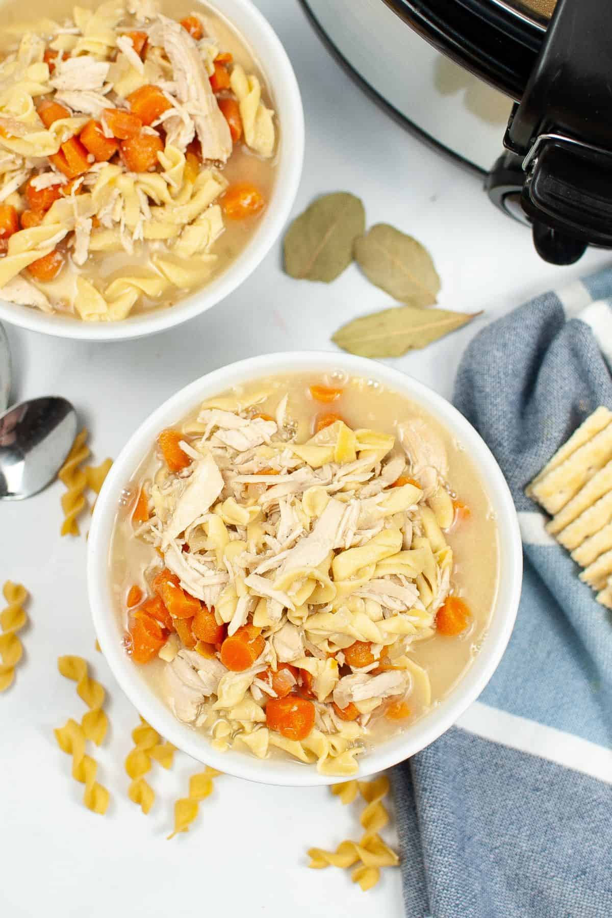 Two bowls of savory Slow Cooker Turkey Noodle Soup with carrots and noodles sit on a white surface near a slow cooker, bay leaves, a spoon, crackers, and uncooked pasta.