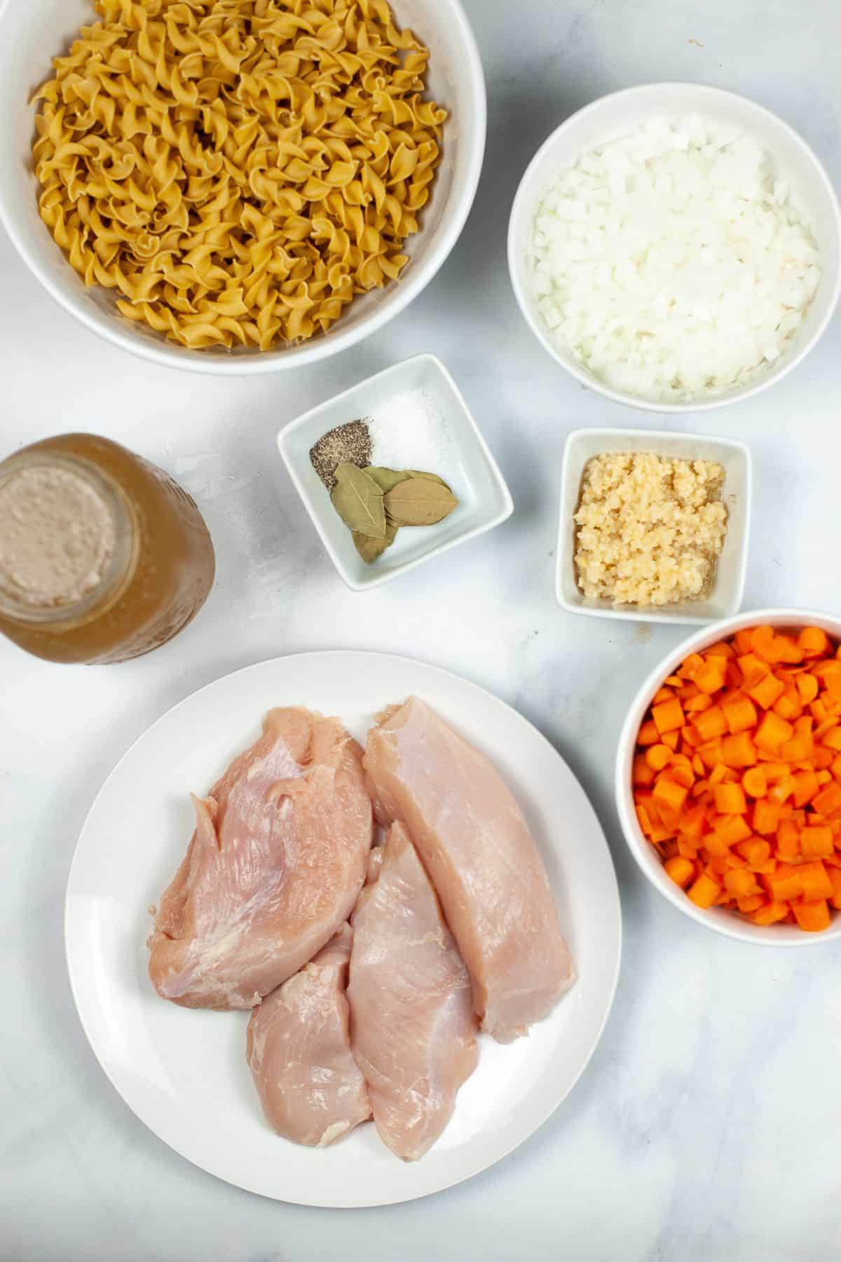 Bowls of uncooked egg noodles, diced onion, chopped carrots, minced garlic, meat, seasonings, and a jar of broth arranged on a white surface.