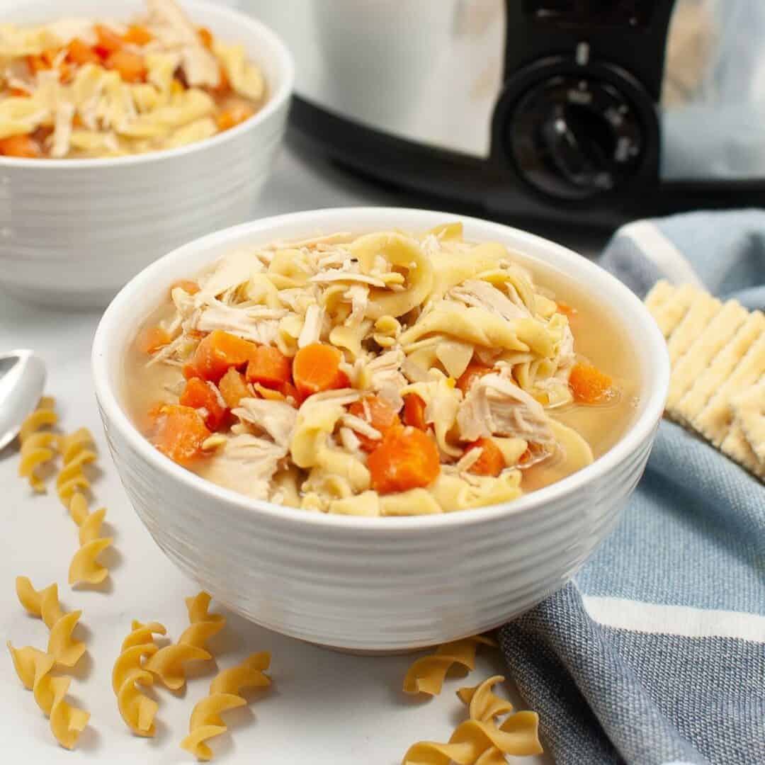 A bowl of chicken noodle soup with carrots and egg noodles sits on a table next to crackers, a blue cloth, and uncooked noodles. In the background, another bowl and a slow cooker suggest Slow Cooker Turkey Noodle Soup is also being served.