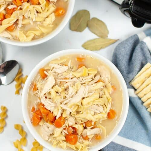 Two bowls of Slow Cooker Turkey Noodle Soup with carrots sit on a white surface next to a slow cooker, bay leaves, a spoon, uncooked pasta, a blue towel, and saltine crackers.
