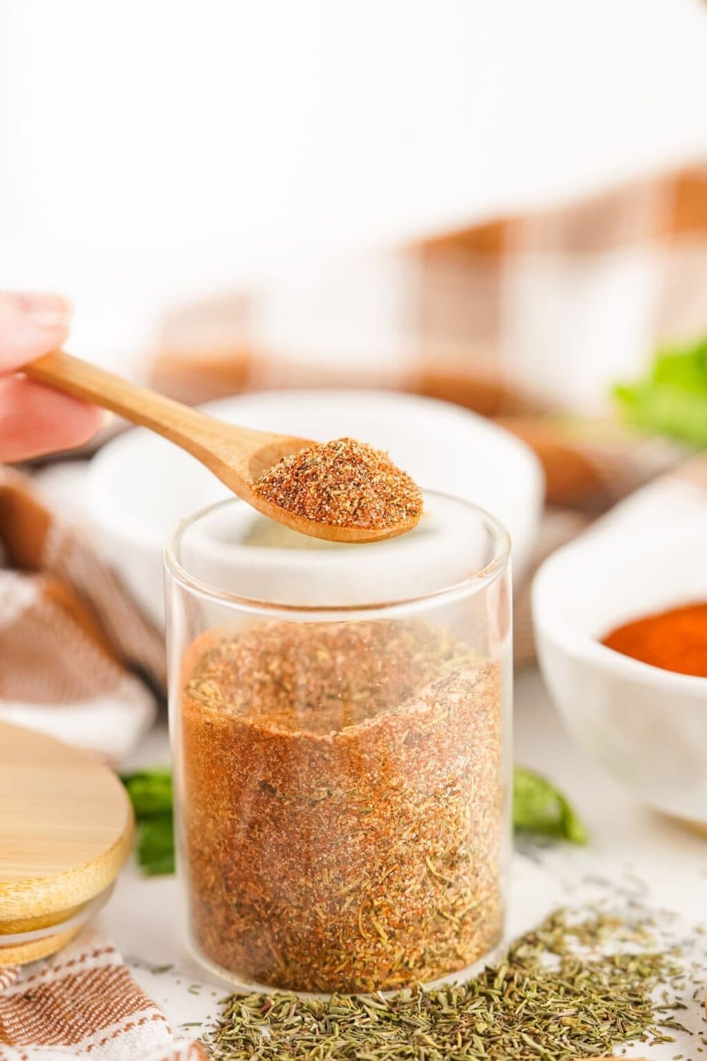 A wooden spoon holds a portion of smoked turkey dry rub above a glass jar filled with the same spice blend, with bowls and herbs in the background.