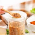 A wooden spoon holds a portion of smoked turkey dry rub above a glass jar filled with the same spice blend, with bowls and herbs in the background.