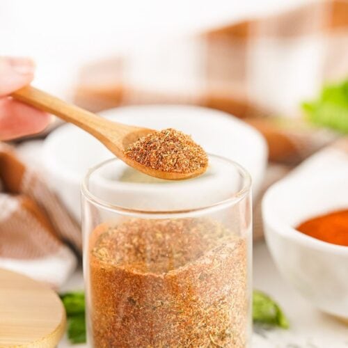 A wooden spoon holds a portion of smoked turkey dry rub above a glass jar filled with the same spice blend, with bowls and herbs in the background.