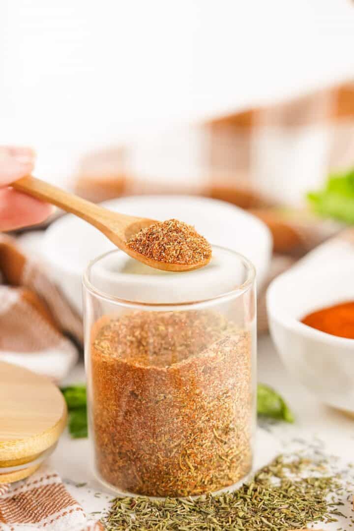A wooden spoon holds a portion of smoked turkey dry rub above a glass jar filled with the same spice blend, with bowls and herbs in the background.