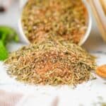A pile of mixed dried herbs and spices, is spilled from a container onto a white surface, with some green leaves in the background.
