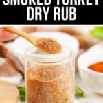 A glass jar filled with smoked turkey dry rub sits on a countertop, with a wooden spoon holding some of the smoked turkey dry rub and bowls of spices in the background.