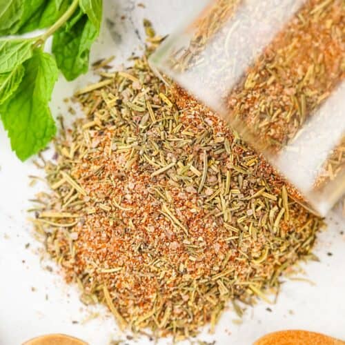 A smoked turkey dry rub spills from a glass jar onto a white surface, with fresh mint leaves and wooden spoons nearby.