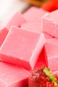 Several pieces of strawberry fudge are stacked closely together, with a fresh strawberry visible in the foreground.