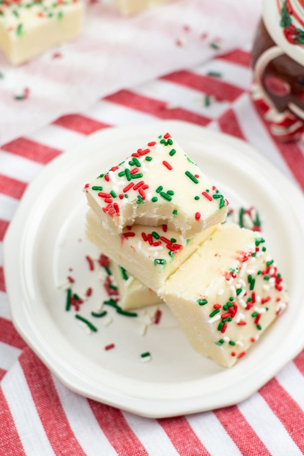 Three pieces of Sugar Cookie Fudge topped with red and green sprinkles are stacked on a white plate, placed on a red and white striped cloth.