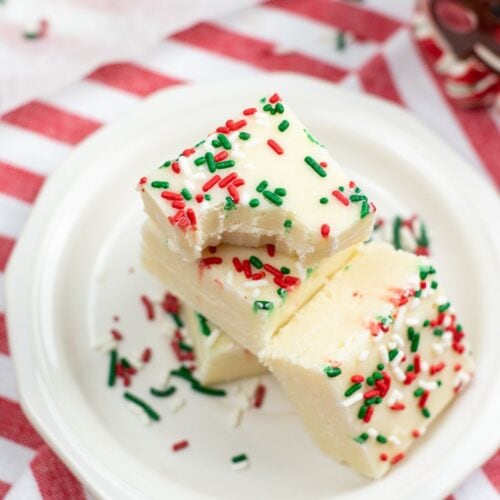 Three pieces of Sugar Cookie Fudge topped with red and green sprinkles are stacked on a white plate, placed on a red and white striped cloth.